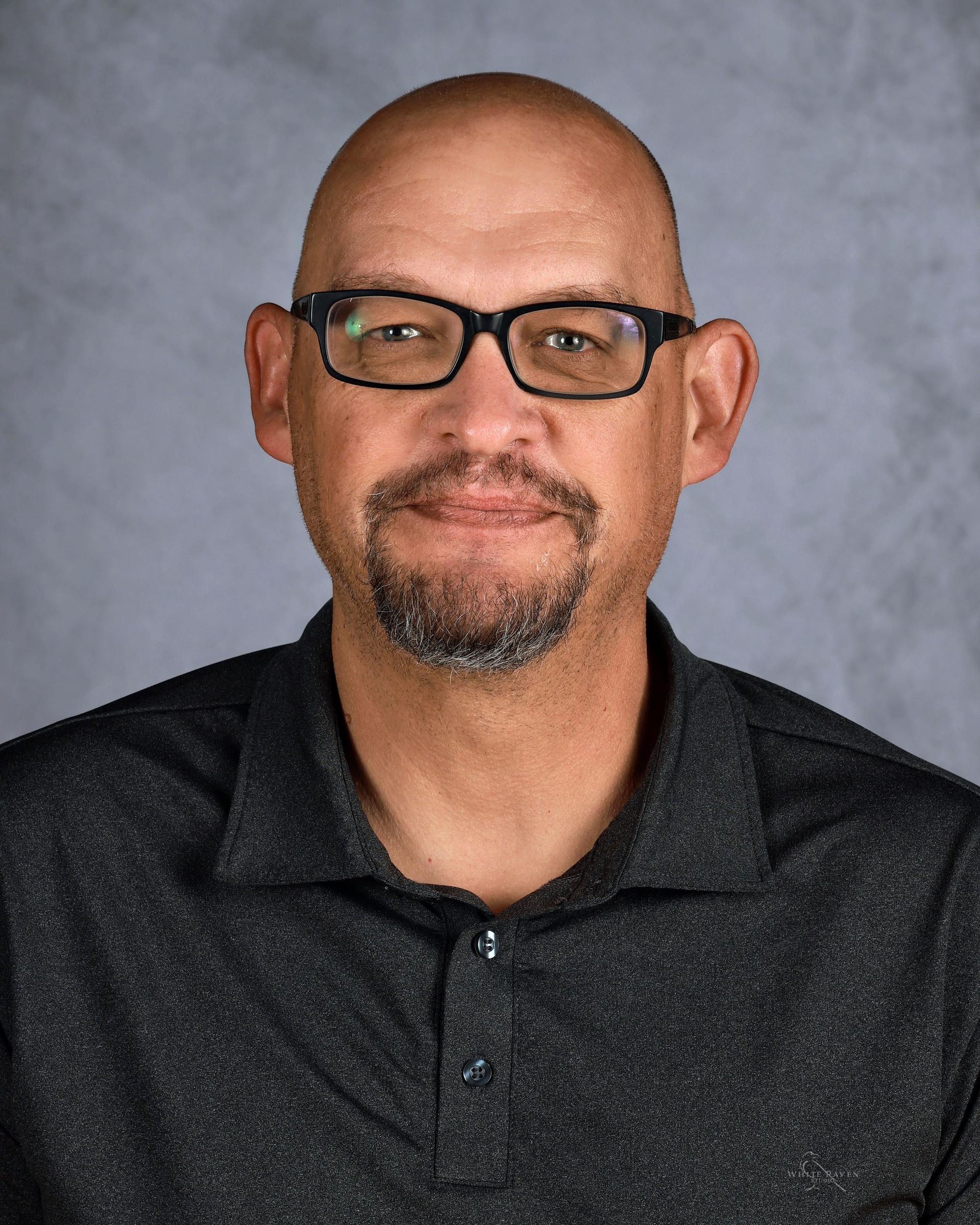 Man wearing glasses and a dark shirt, smiling, with a short beard, in front of a gray backdrop.