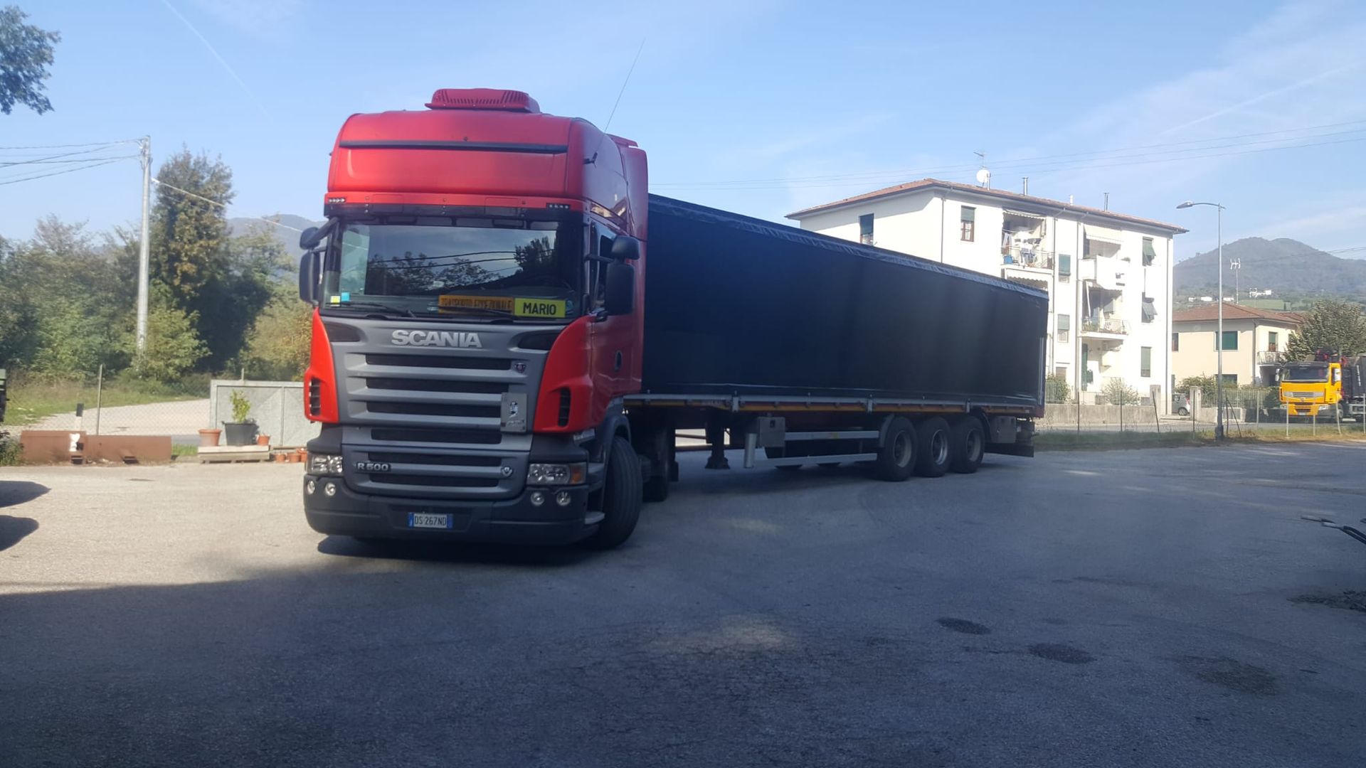 Un autoarticolato Scania rosso con un rimorchio nero parcheggiato su un piazzale asfaltato in una giornata di sole.