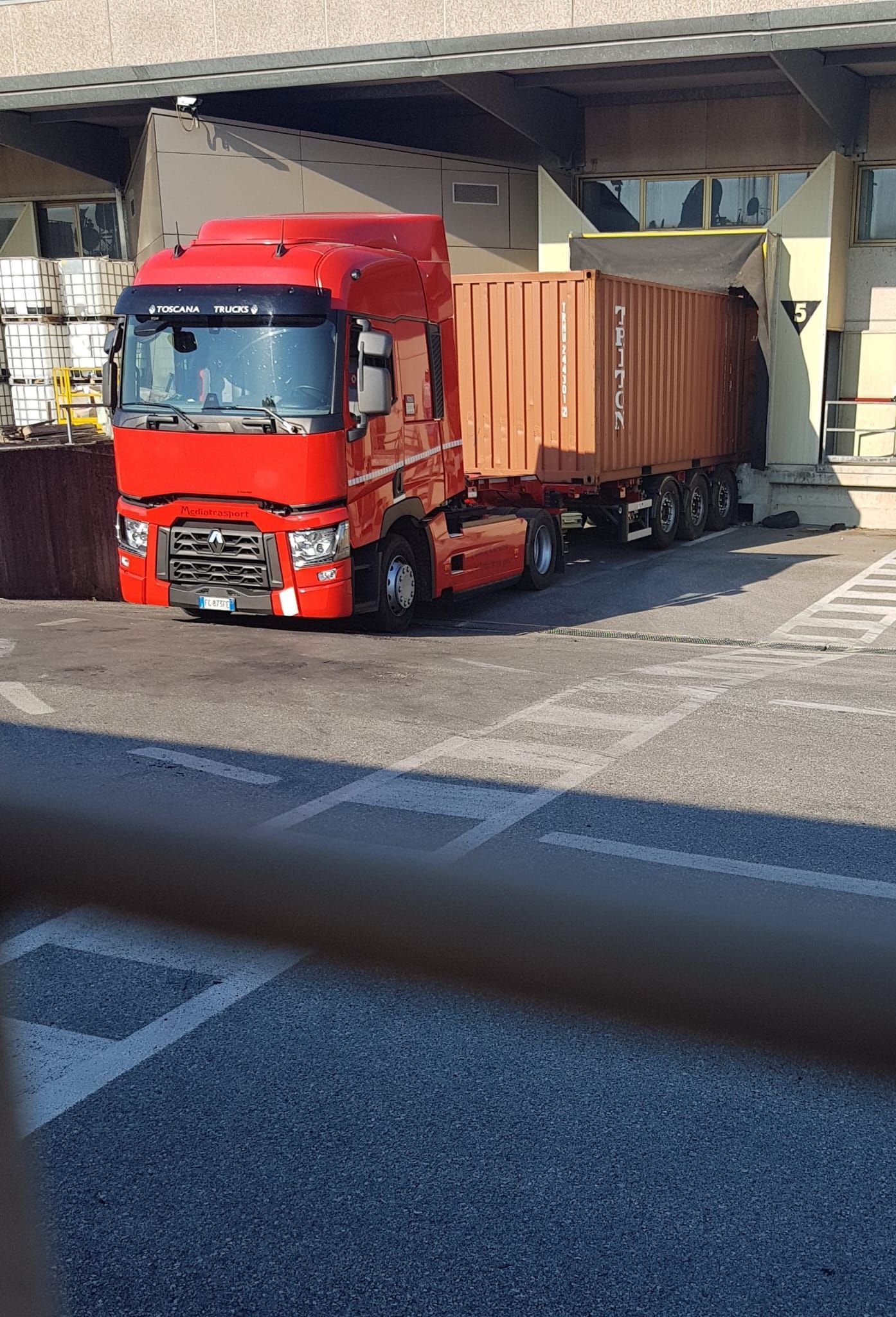 Camion rosso con rimorchio parcheggiato alla banchina di carico. Il container è marrone chiaro, inquadratura esterna.