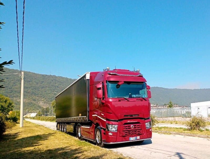 Un semirimorchio rosso con rimorchio percorre una strada con verdi colline sullo sfondo e un cielo azzurro.