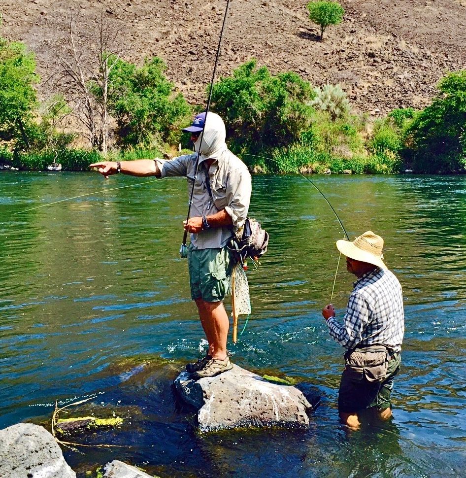 Two men are fishing in a river and one is standing on a rock