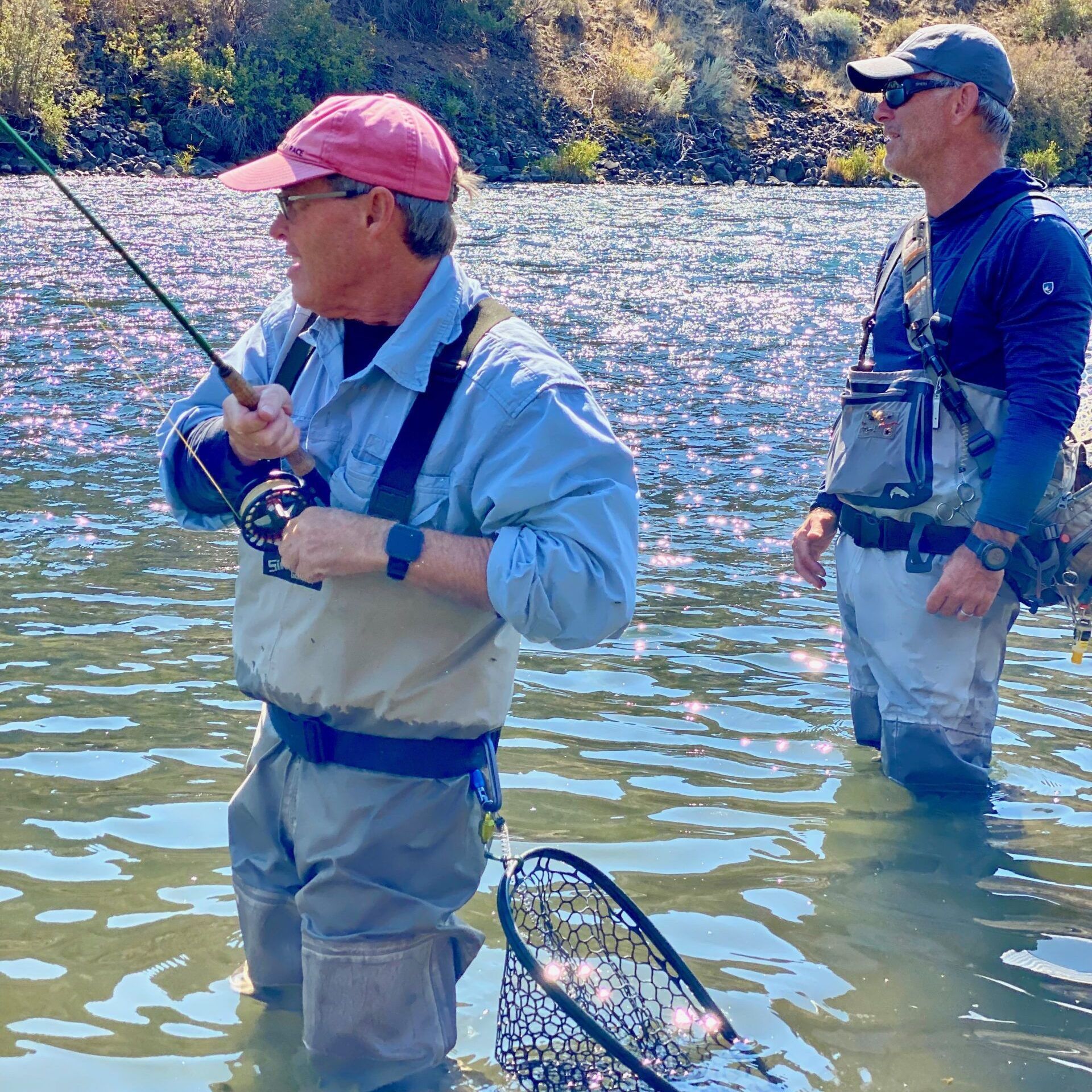 Two men are fishing in a river and one is wearing a pink hat