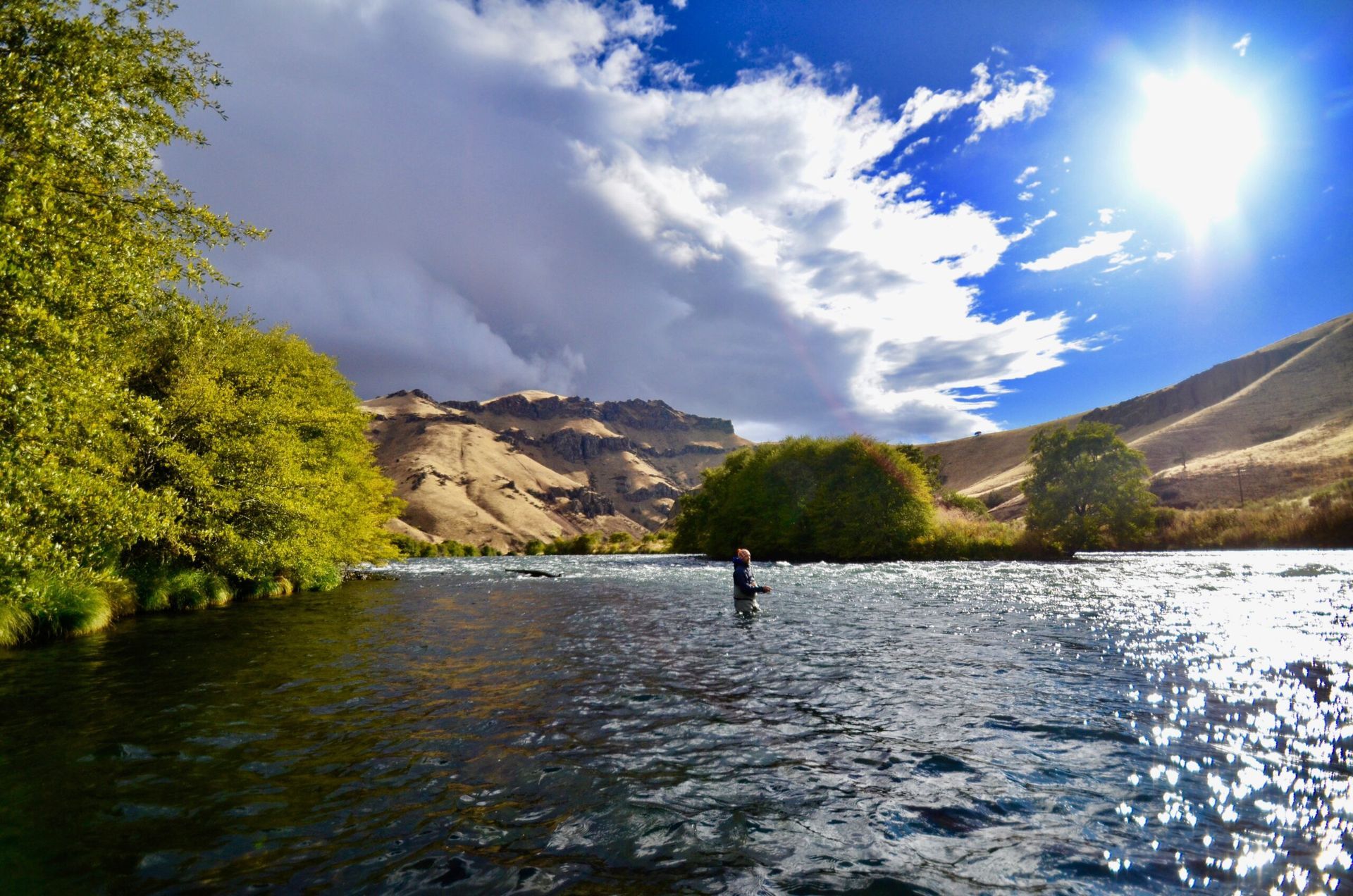 A man is fishing in a river with mountains in the background.