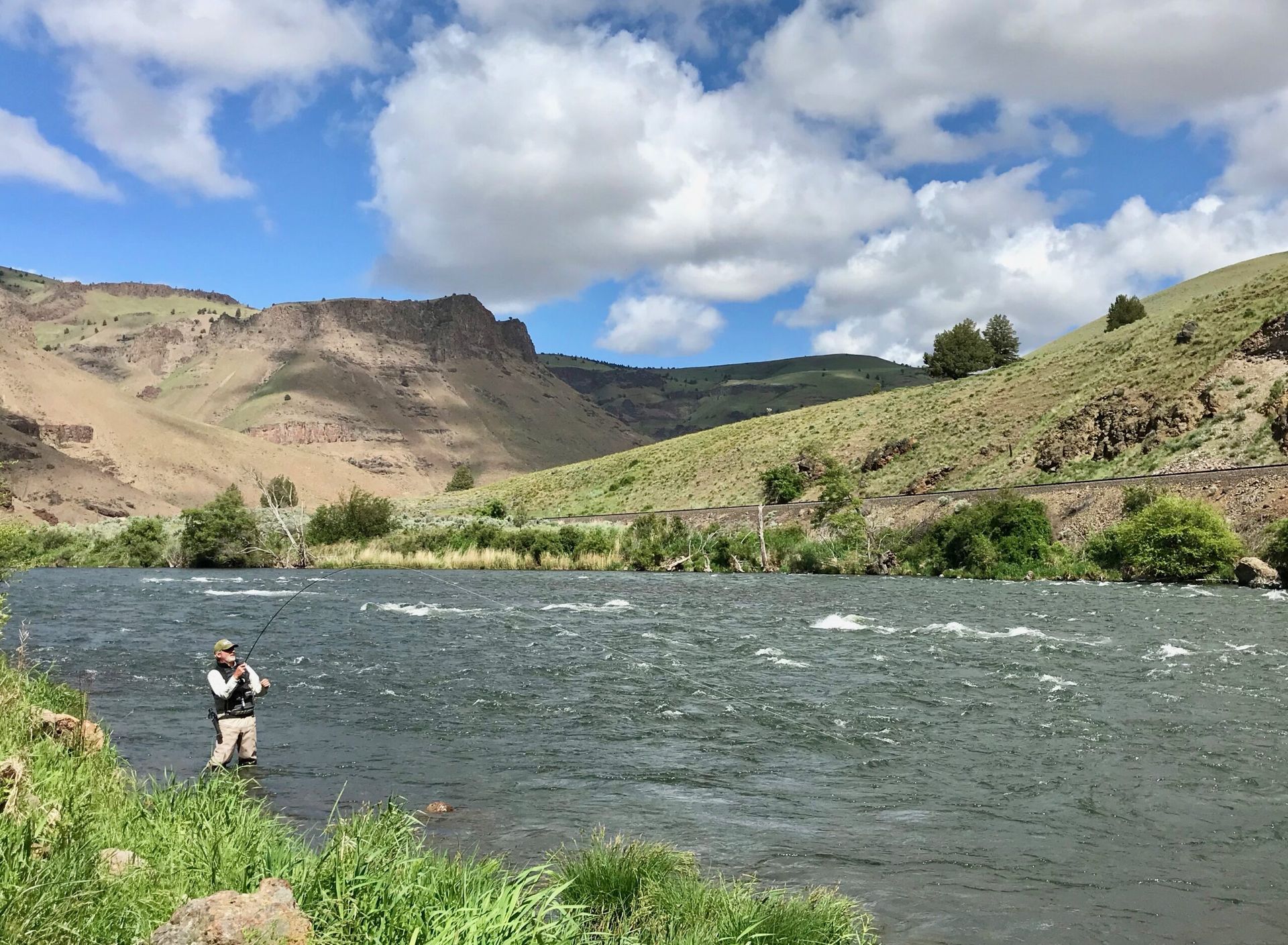 A man is fishing in a river with mountains in the background