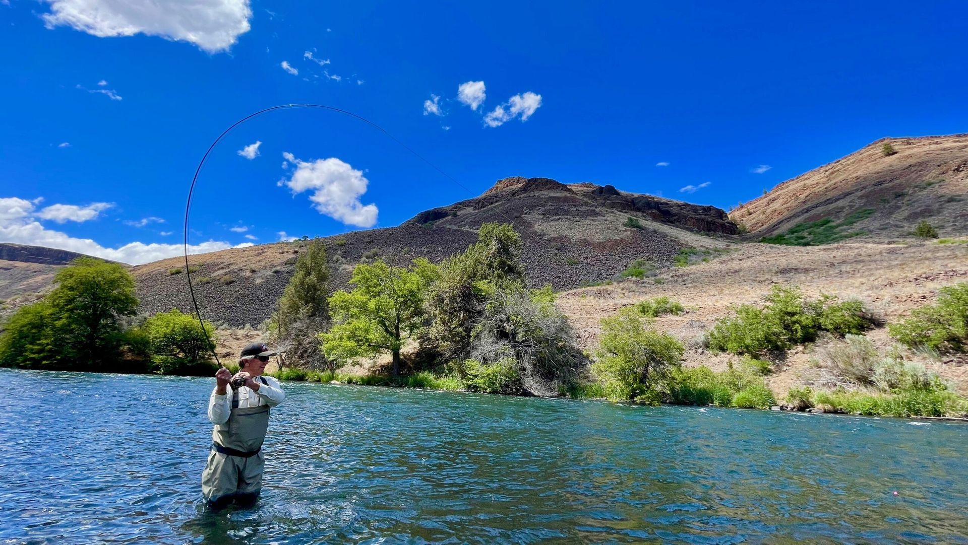 A man is fishing in a river with mountains in the background.