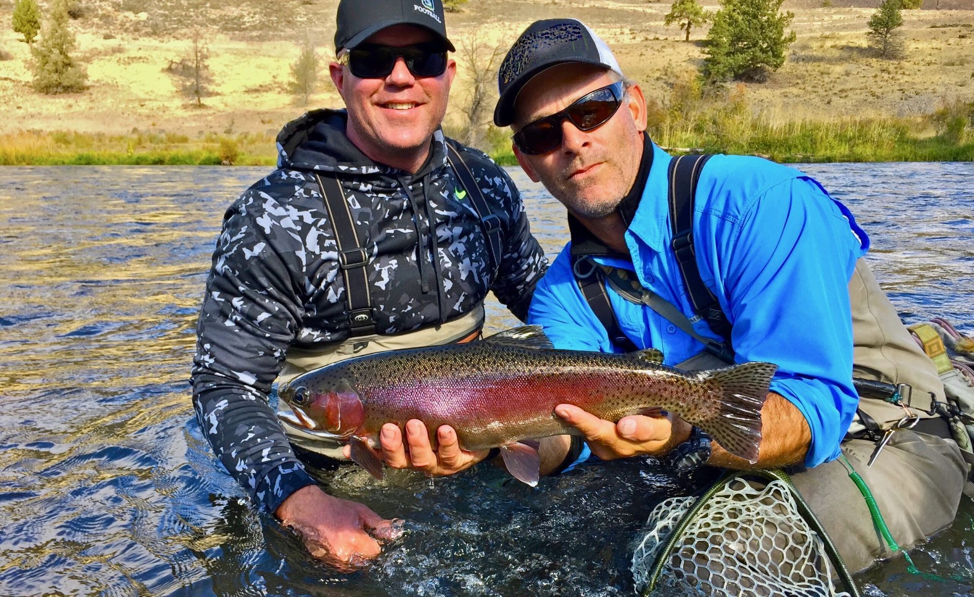 Two men are holding a large rainbow trout in a river.