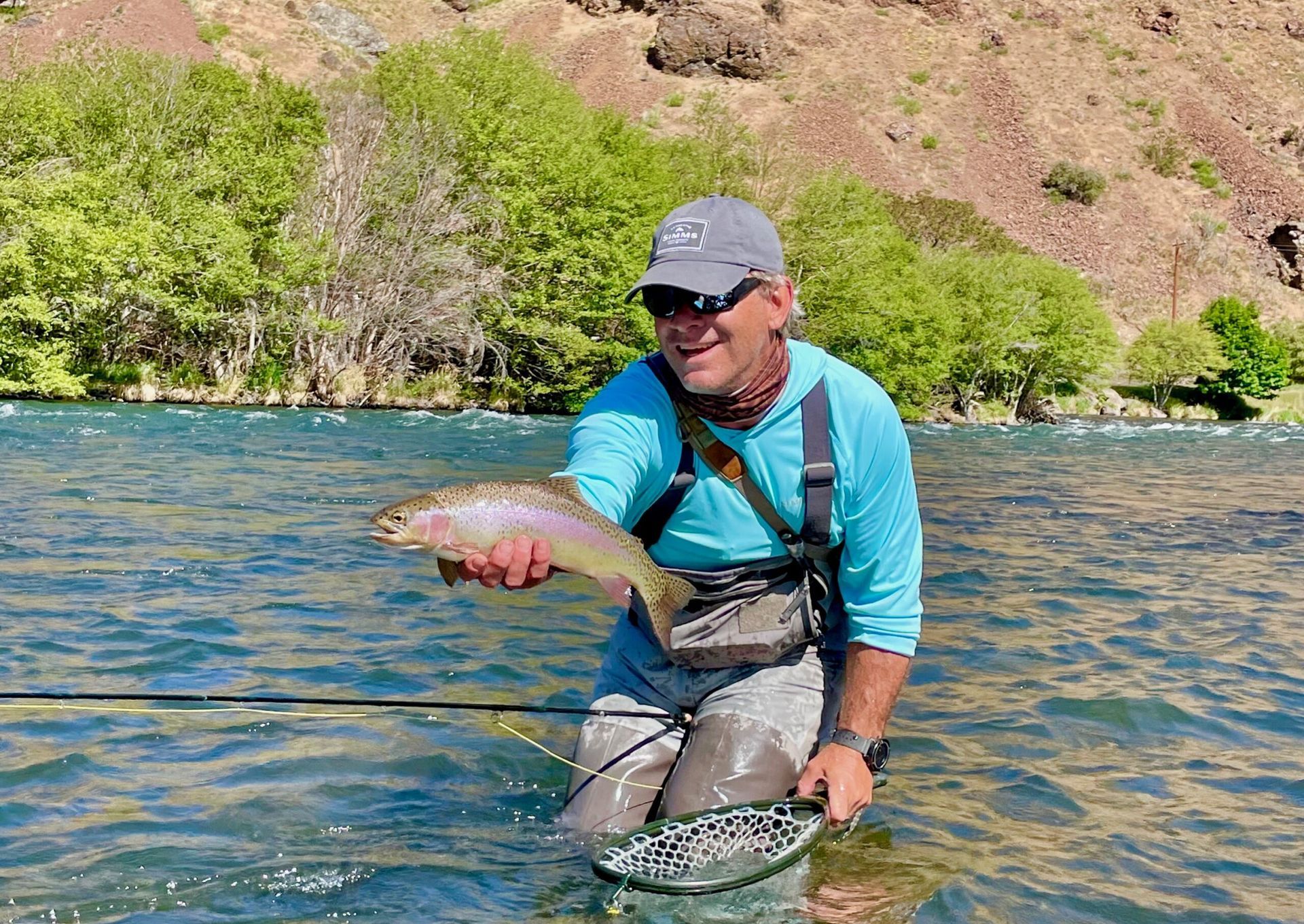 A man is fishing in a river and holding a rainbow trout.