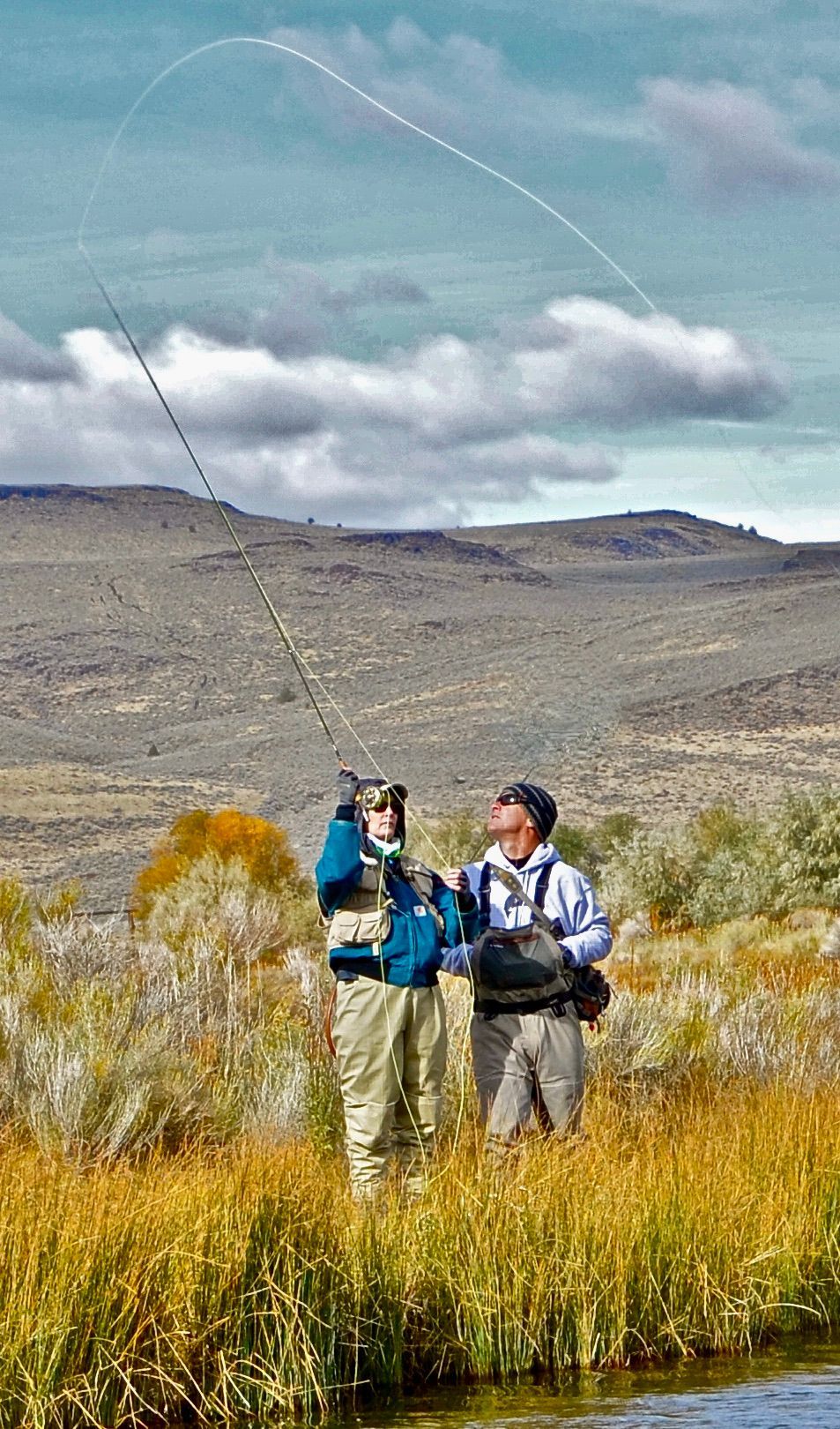 Two men are fishing in a river with mountains in the background.