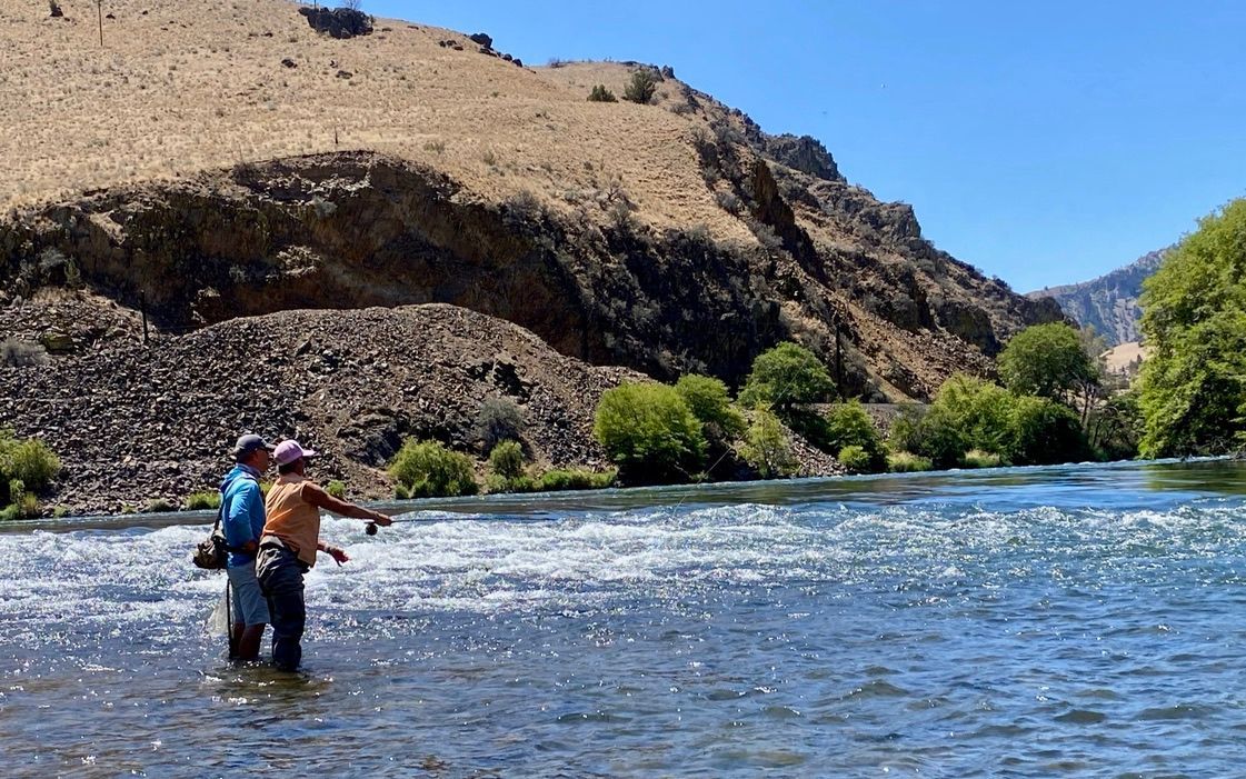 Two men are fishing in a river with mountains in the background