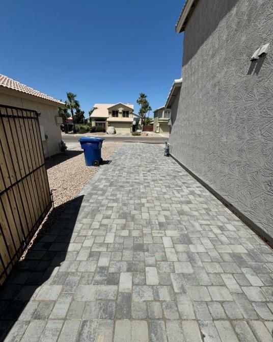 A brick walkway leading to a house with a blue trash can on the side