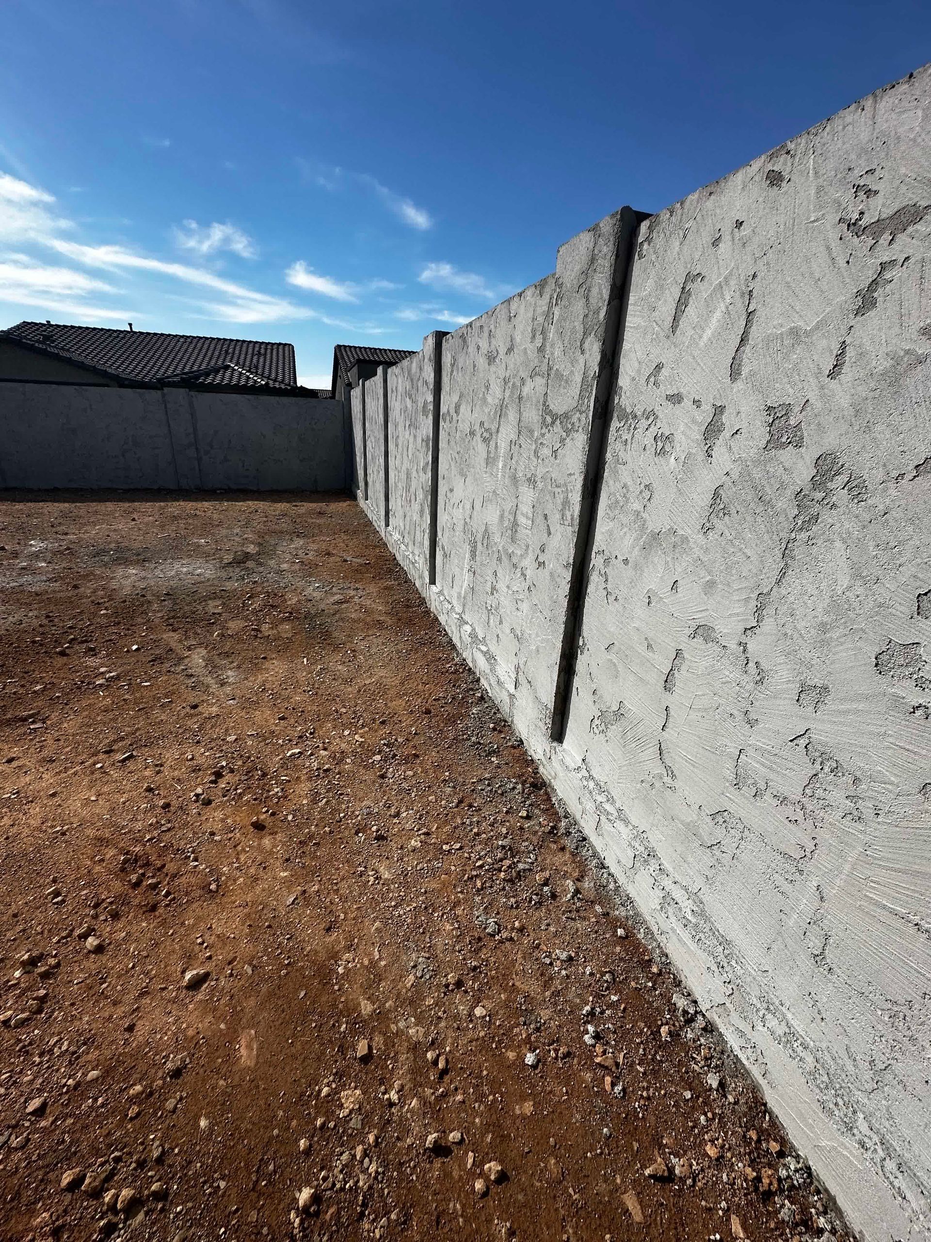 A concrete fence surrounds a dirt field with a house in the background.