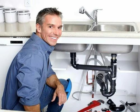 A plumber is kneeling under a sink in a kitchen.