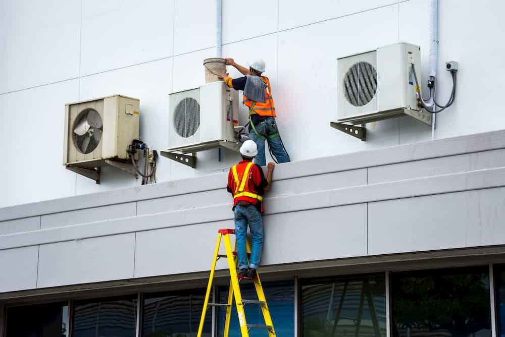 Two men are working on air conditioners on the side of a building.