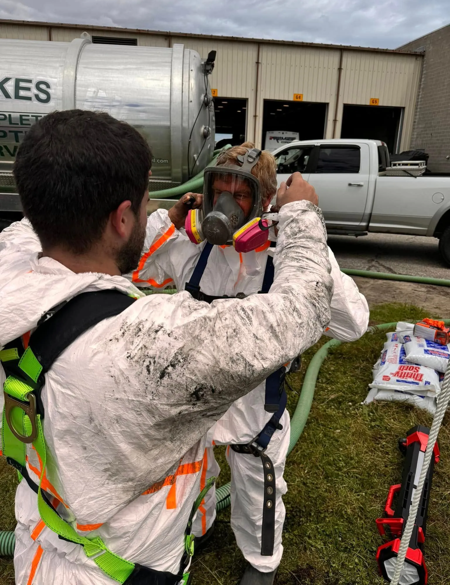 Two people in hazmat suits, one assisting the other with a respirator. Outdoors, near a truck.
