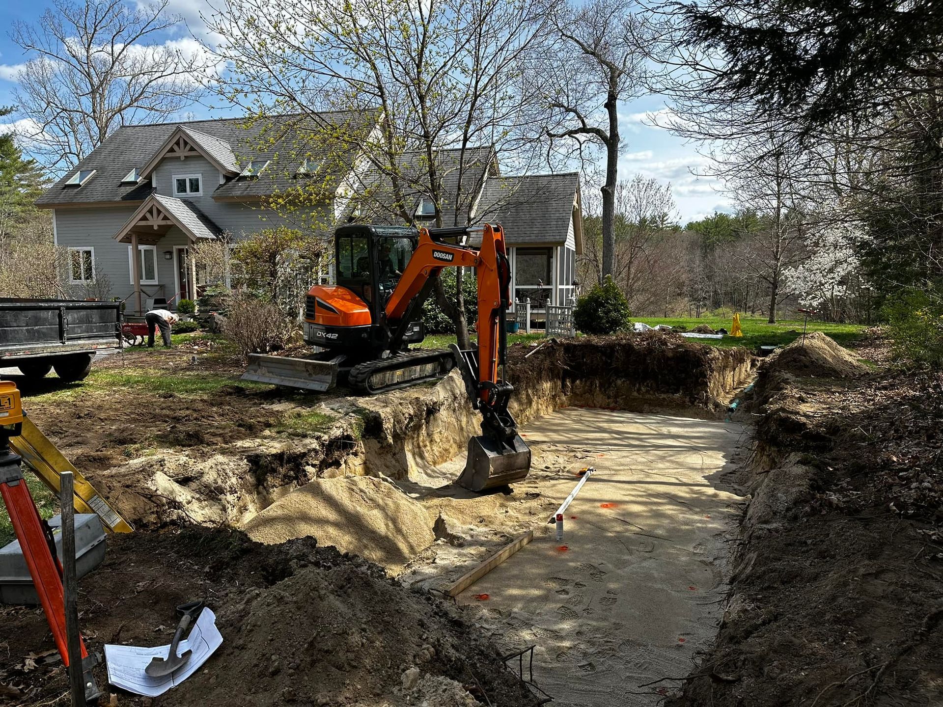 Excavator digging a rectangular foundation in a residential backyard with a house visible in the background.