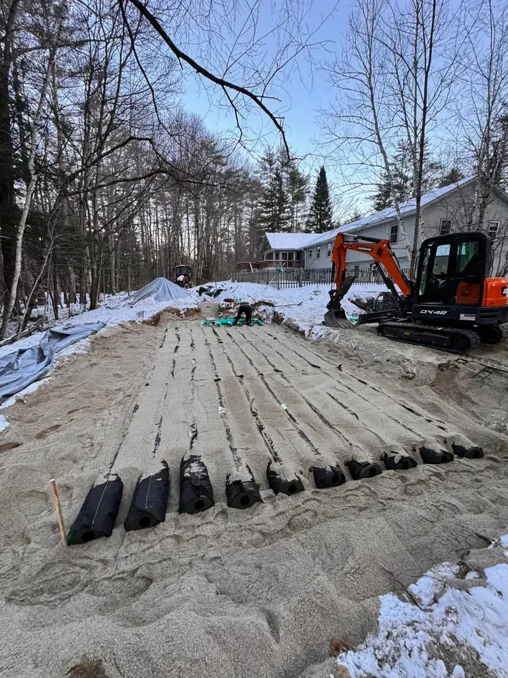 An excavator on a construction site with wood beams laid out, covered in sand, in a snowy, wooded area.