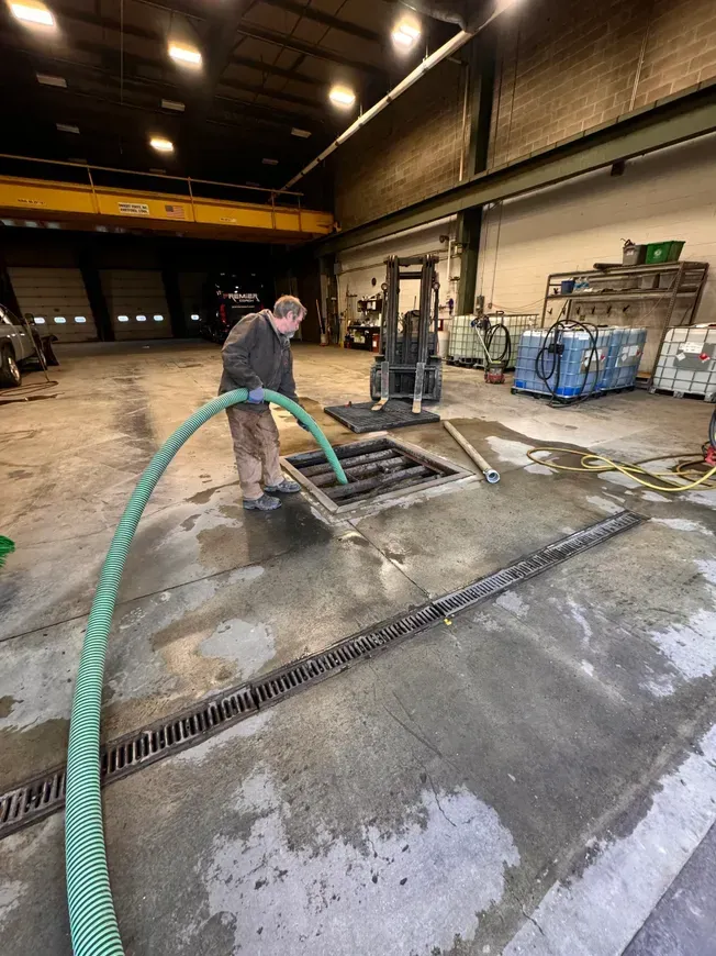Man using a hose to clean a drain in a warehouse with a forklift in the background.