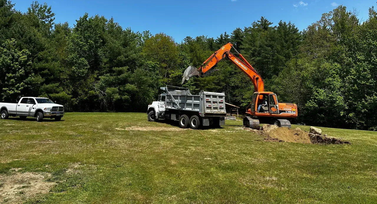 An excavator loading dirt into a dump truck; a white pickup truck is parked nearby on a grassy field with trees.
