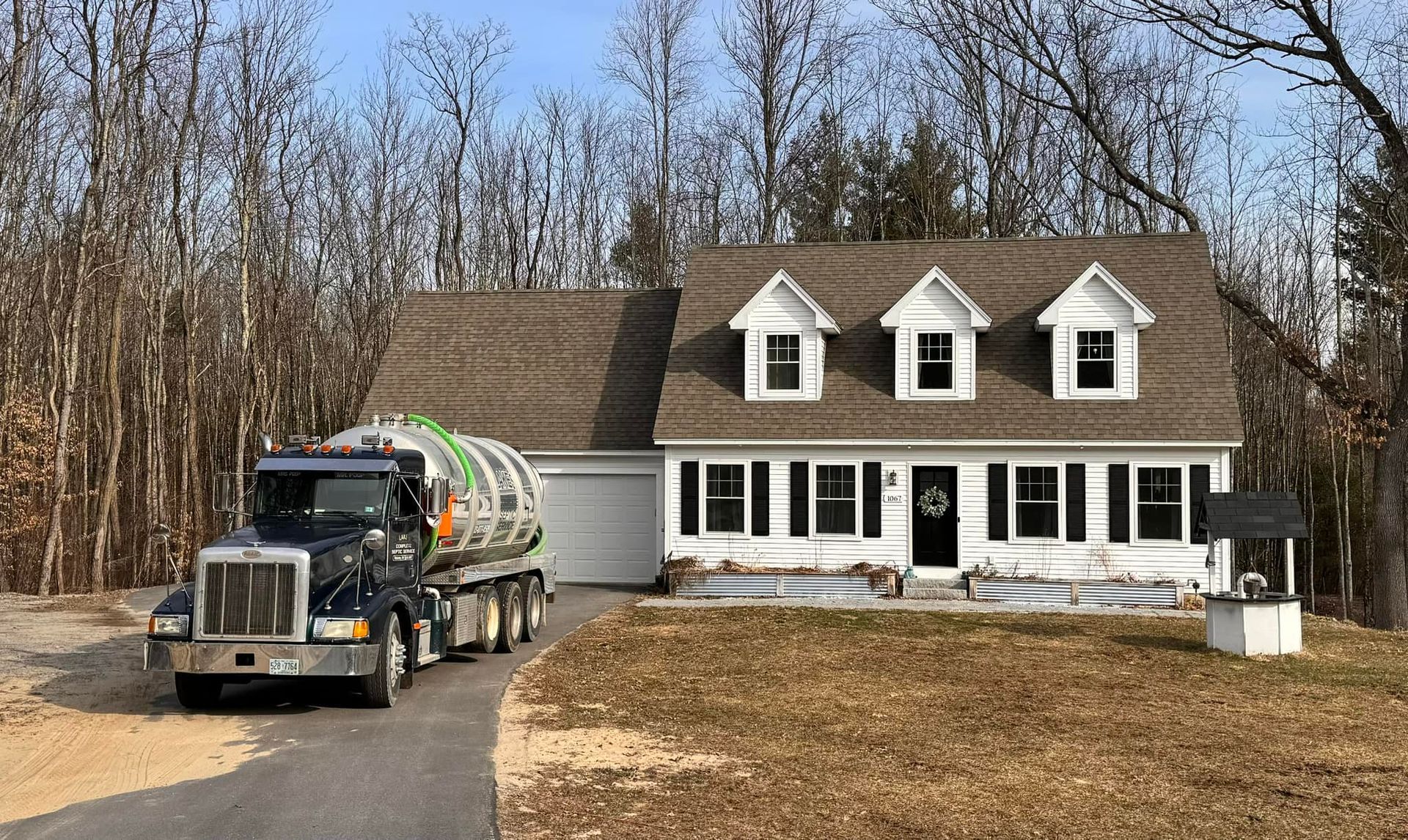 Septic truck parked in front of a white house with black shutters and a brown roof. Well in yard.