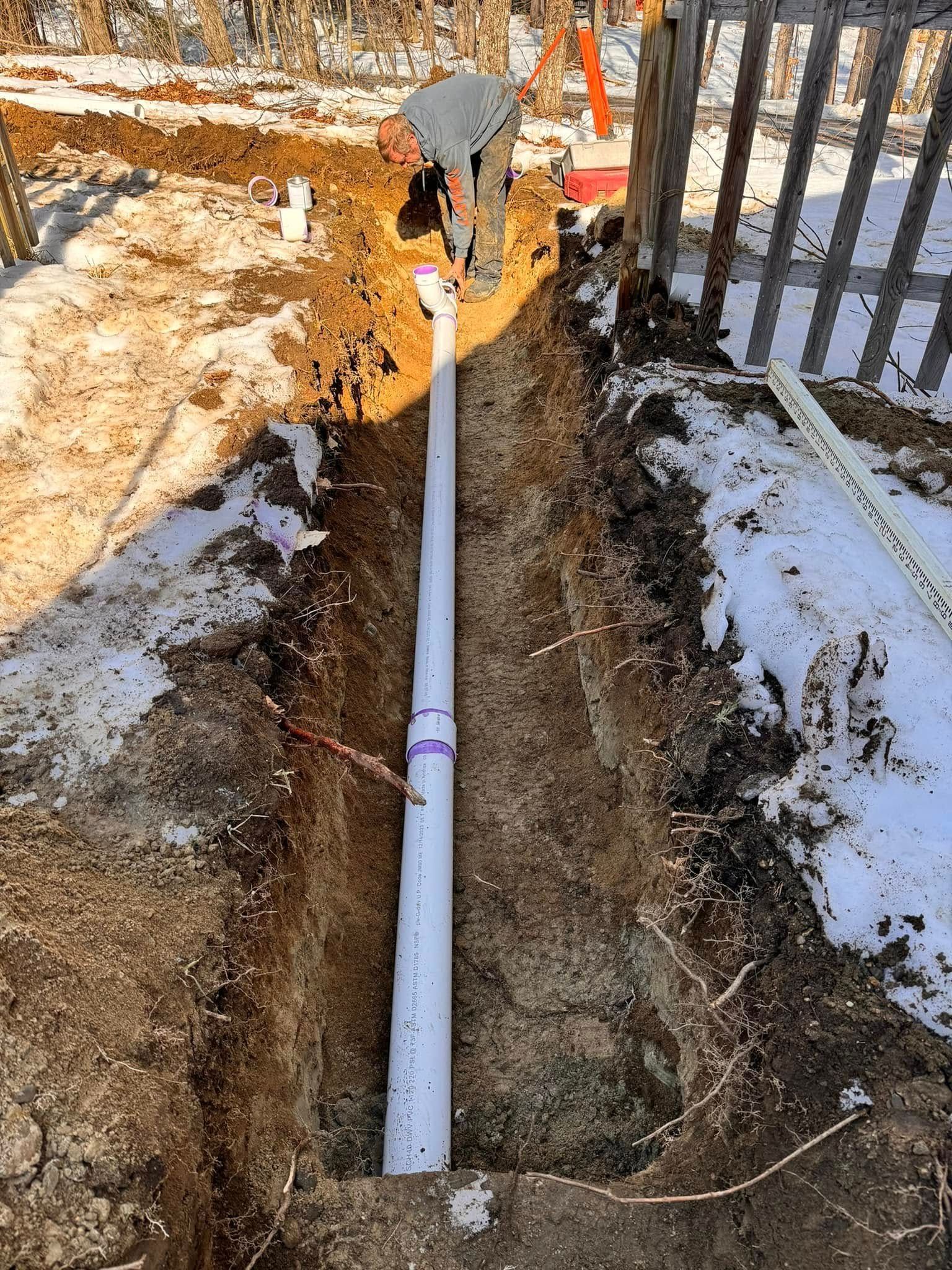 A long white PVC pipe in a trench, likely for drainage, next to a weathered wooden fence and snow.