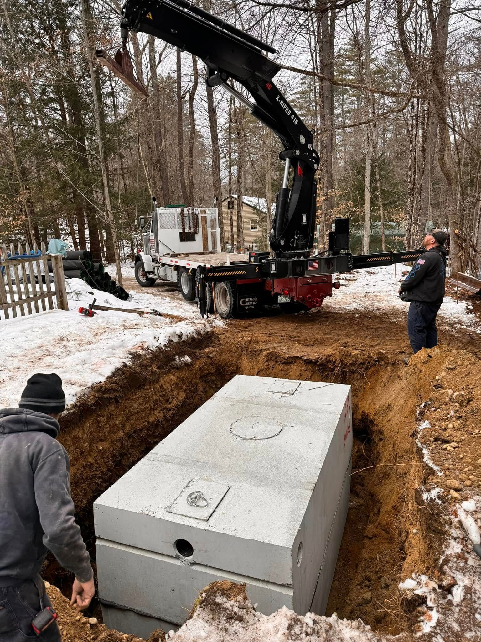 A concrete septic tank being lowered into an excavation by a crane in a snowy yard. Two men watch.