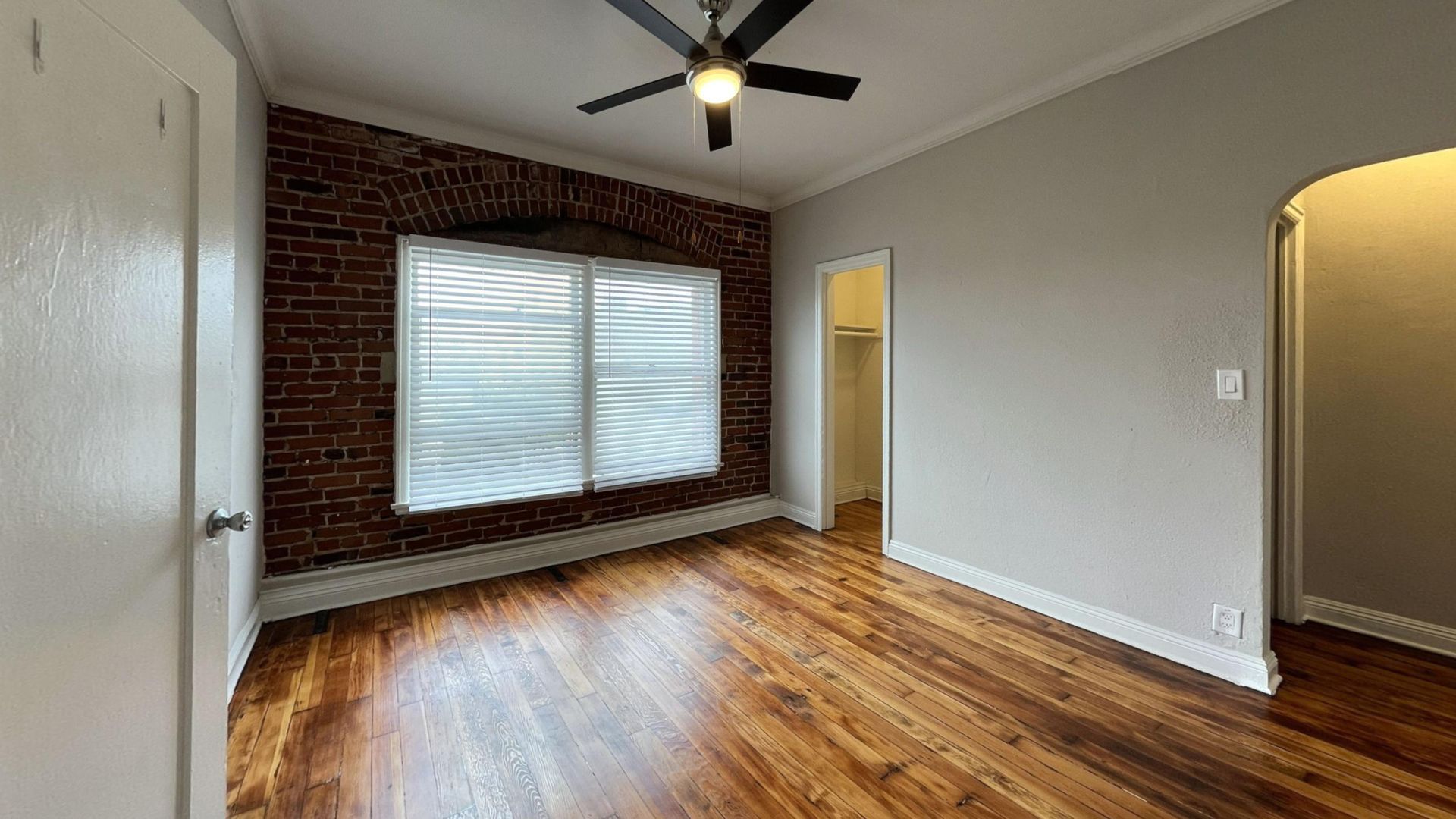 Empty bedroom with exposed brick wall, window, wooden floor, and ceiling fan.