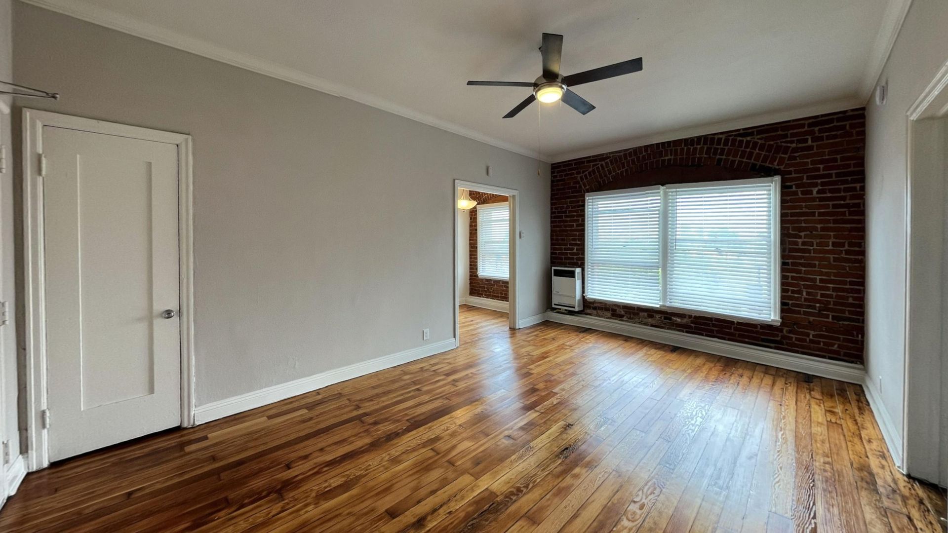 Living room with hardwood floors, exposed brick wall, white door, and a ceiling fan.