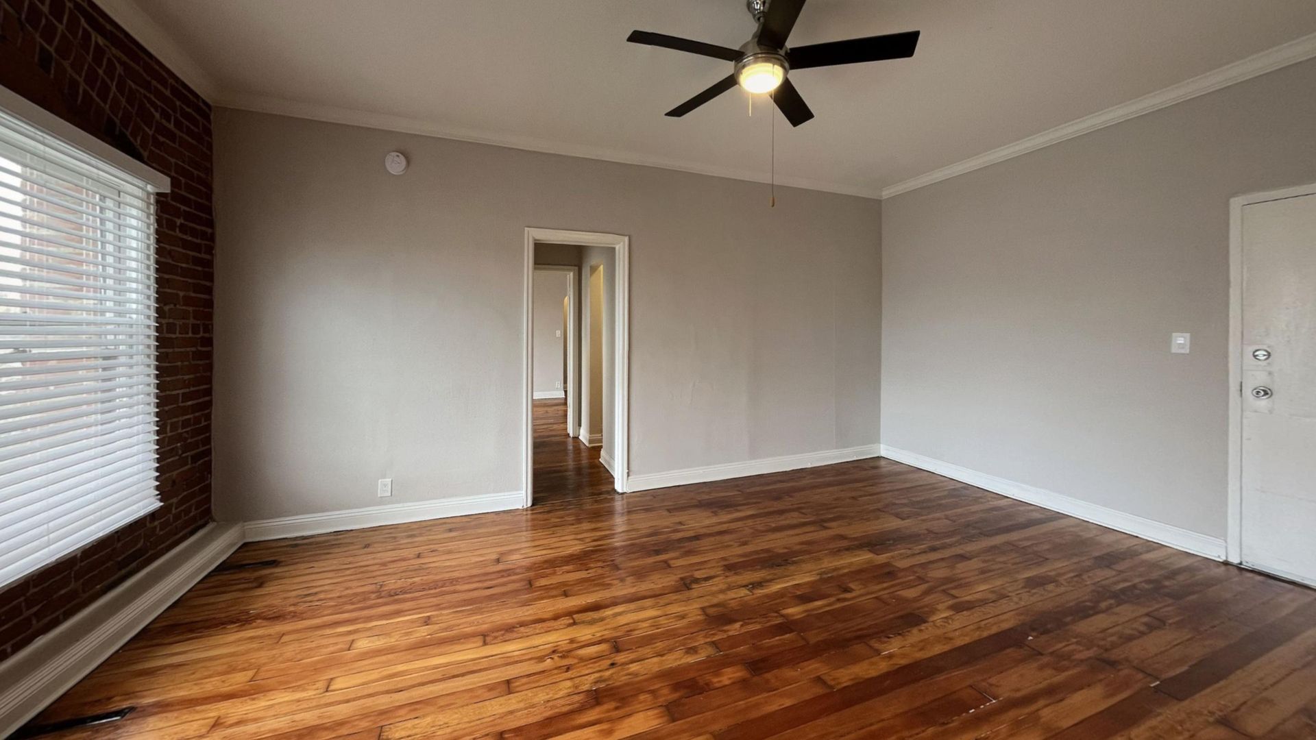 Empty room with hardwood floors, exposed brick wall, and a ceiling fan.