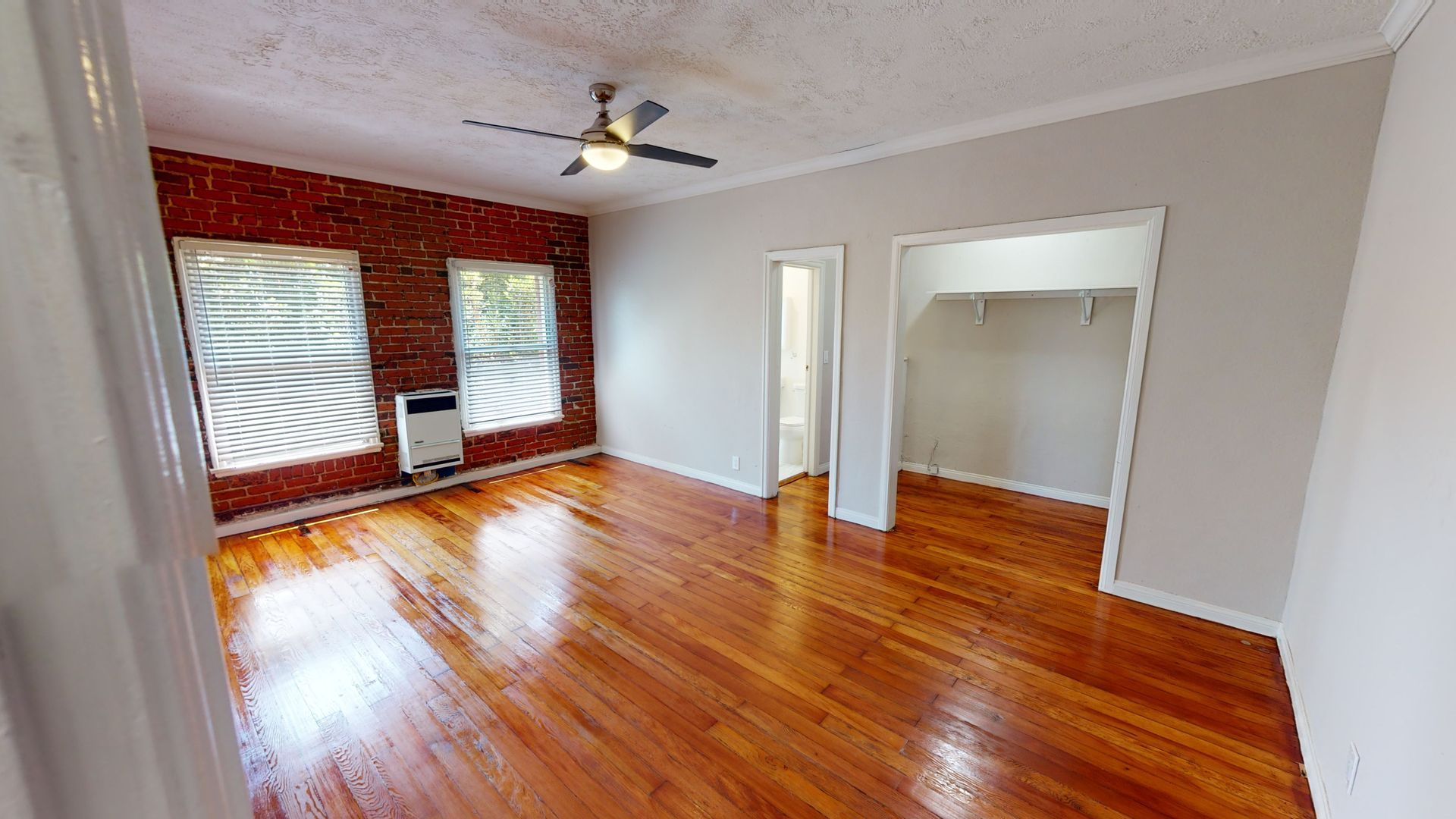 Empty room with hardwood floors, exposed brick wall, windows, ceiling fan, and open doorways.