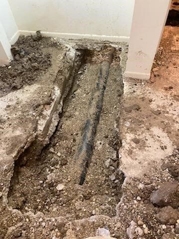 Trench dug in floor exposing two pipes, likely for plumbing repair. Interior, dirt, debris.
