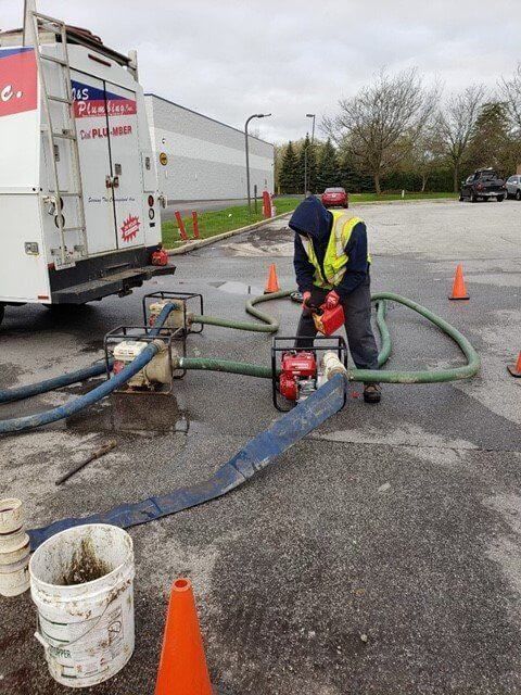 Man in safety vest operating a red pump with hoses, likely draining something. Near a truck and cones.