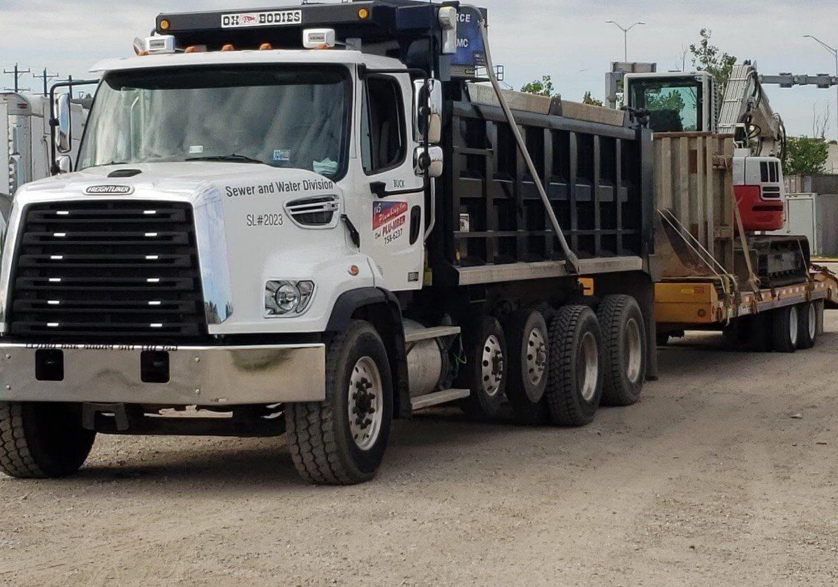 White dump truck with excavator on trailer, parked on gravel.
