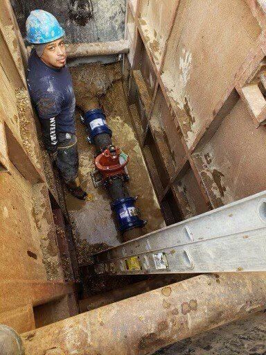 Man in hard hat working on pipes in a trench. Blue and red valves. Ladder inside.
