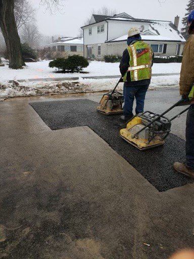 Two workers compacting asphalt patch on a snowy road.