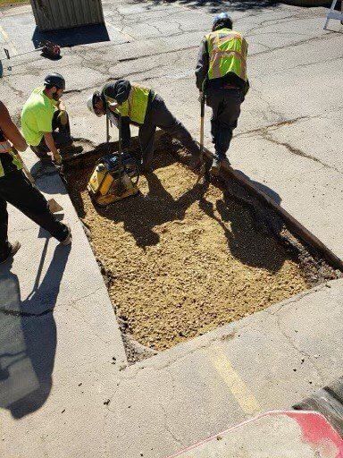 Road workers repairing asphalt: three in vests and hard hats, one using a compactor, one shoveling.