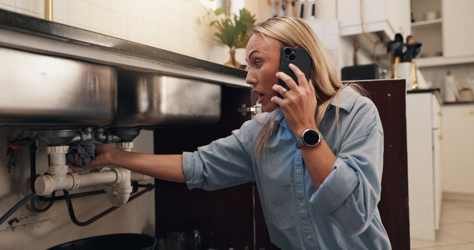 A woman inspecting pipes under a kitchen sink while using the phone to report the plumbing issue A woman inspecting pipes under a kitchen sink while using the phone to report the plumbing issue