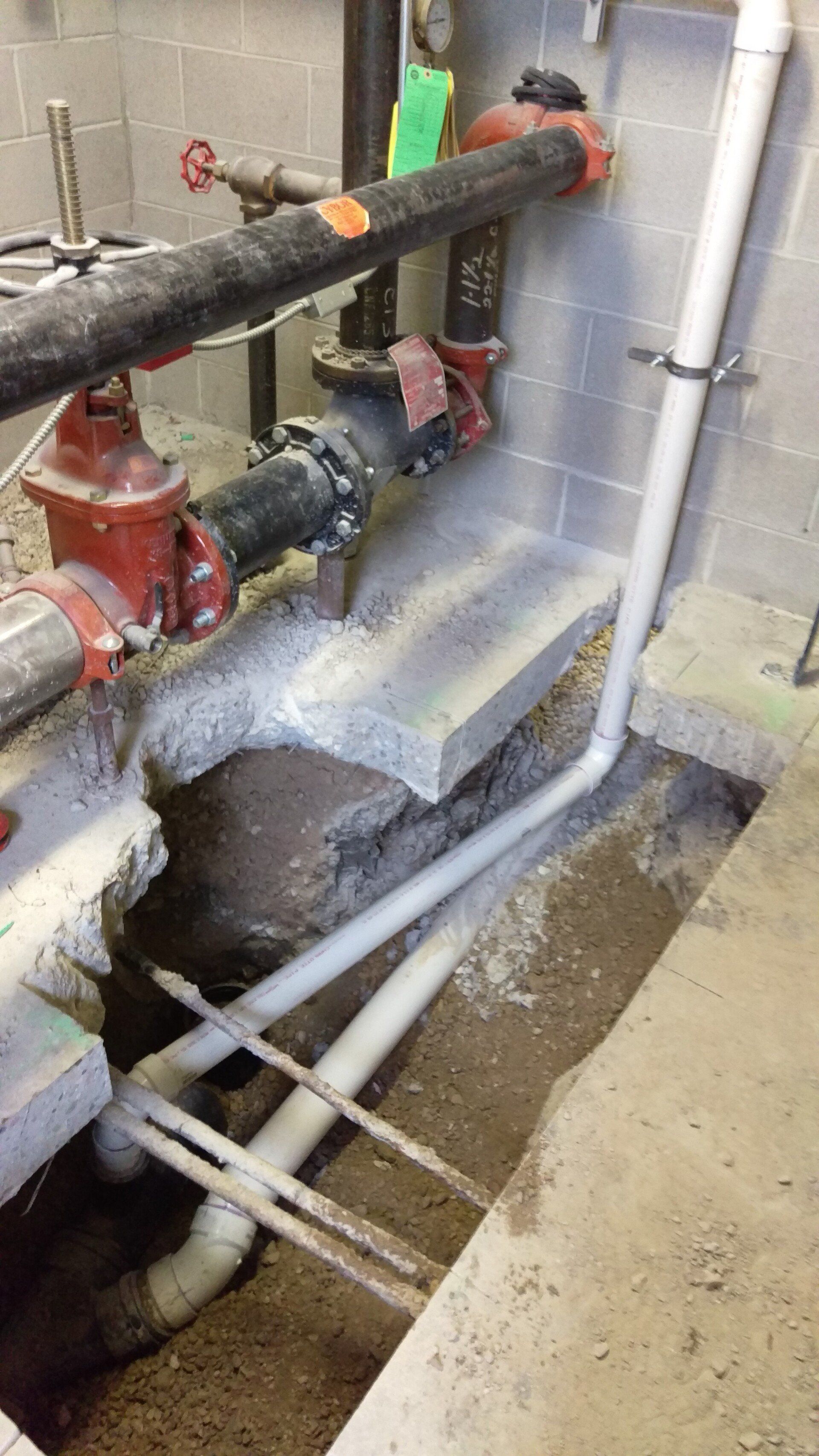 Pipes in a trench, near a concrete wall with valves and plumbing components.