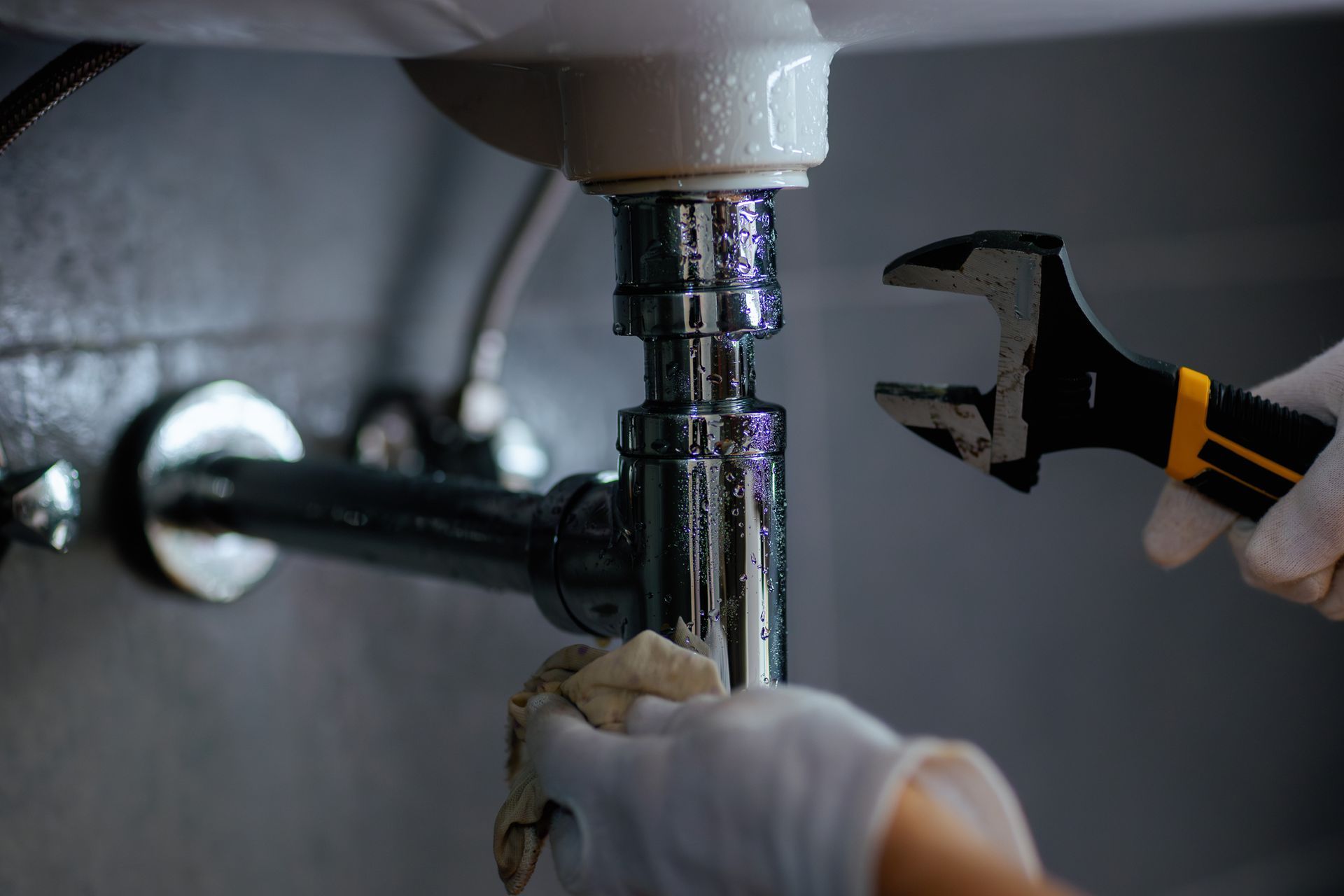 Close-up of a plumber with white gloves as they use a wrench to work on a sink’s pipes.