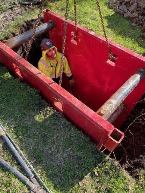 Construction worker in yellow jacket inside a red trench box in a dirt pit, with metal pipes visible.