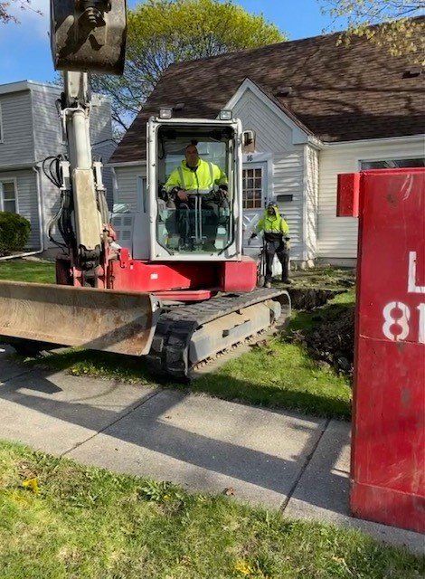 An excavator digging near a house; two workers in vests, one operating the machine.