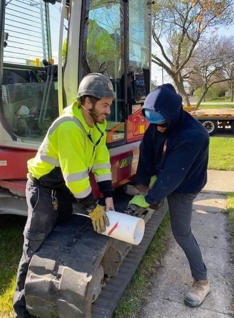 Two construction workers in safety gear assemble a pipe near a small excavator outdoors.