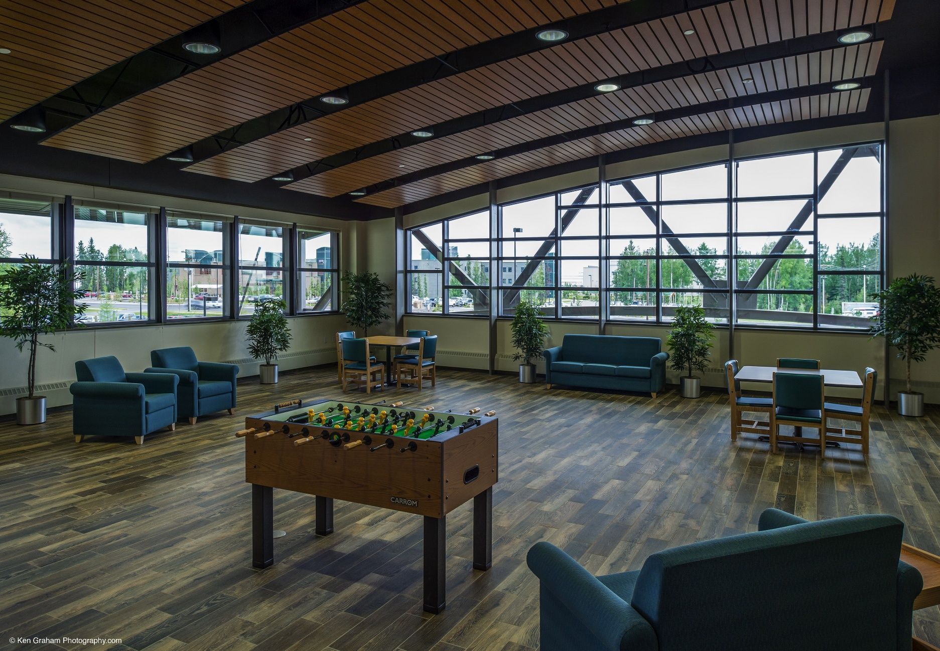 A brightly lit common room with seating, tables, and a foosball table. Large windows offer an outside view.