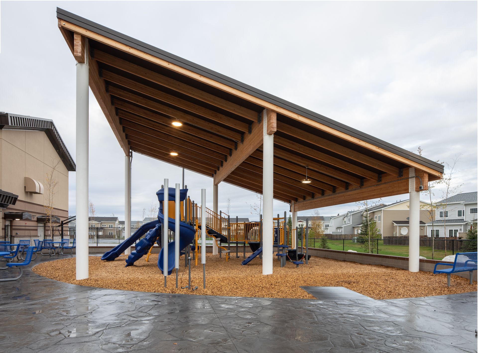 Playground with wood-beamed shelter; slides, wood chips, and blue play structures. Overcast sky.