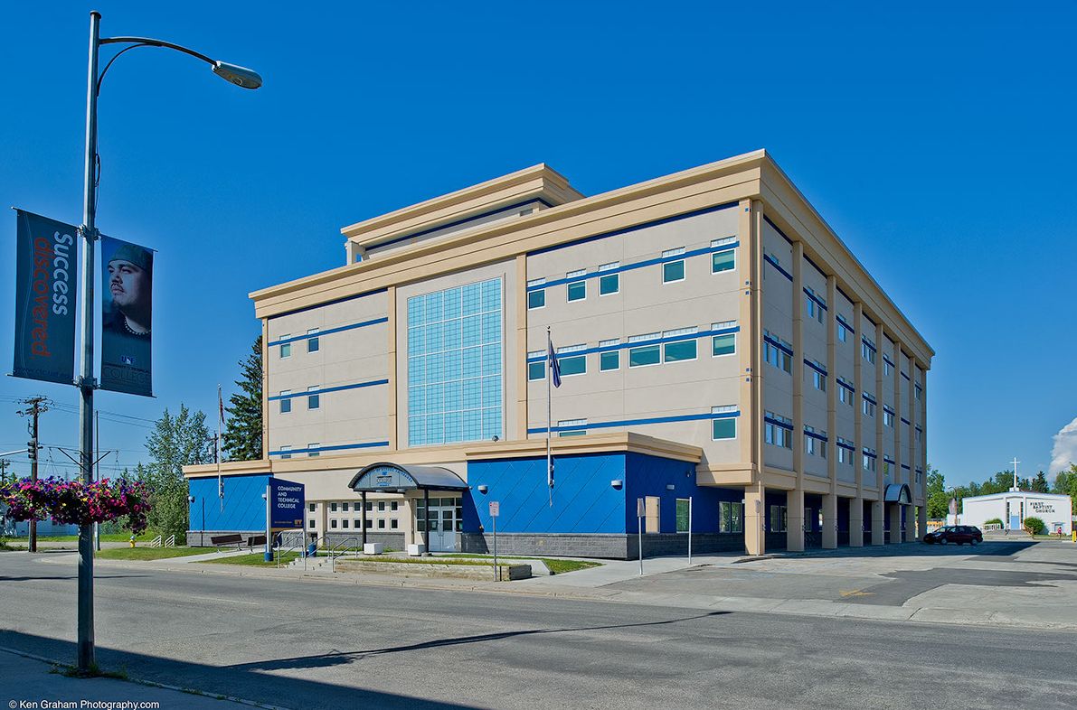 Modern beige and blue building with large windows under a clear blue sky; sign and street in front.