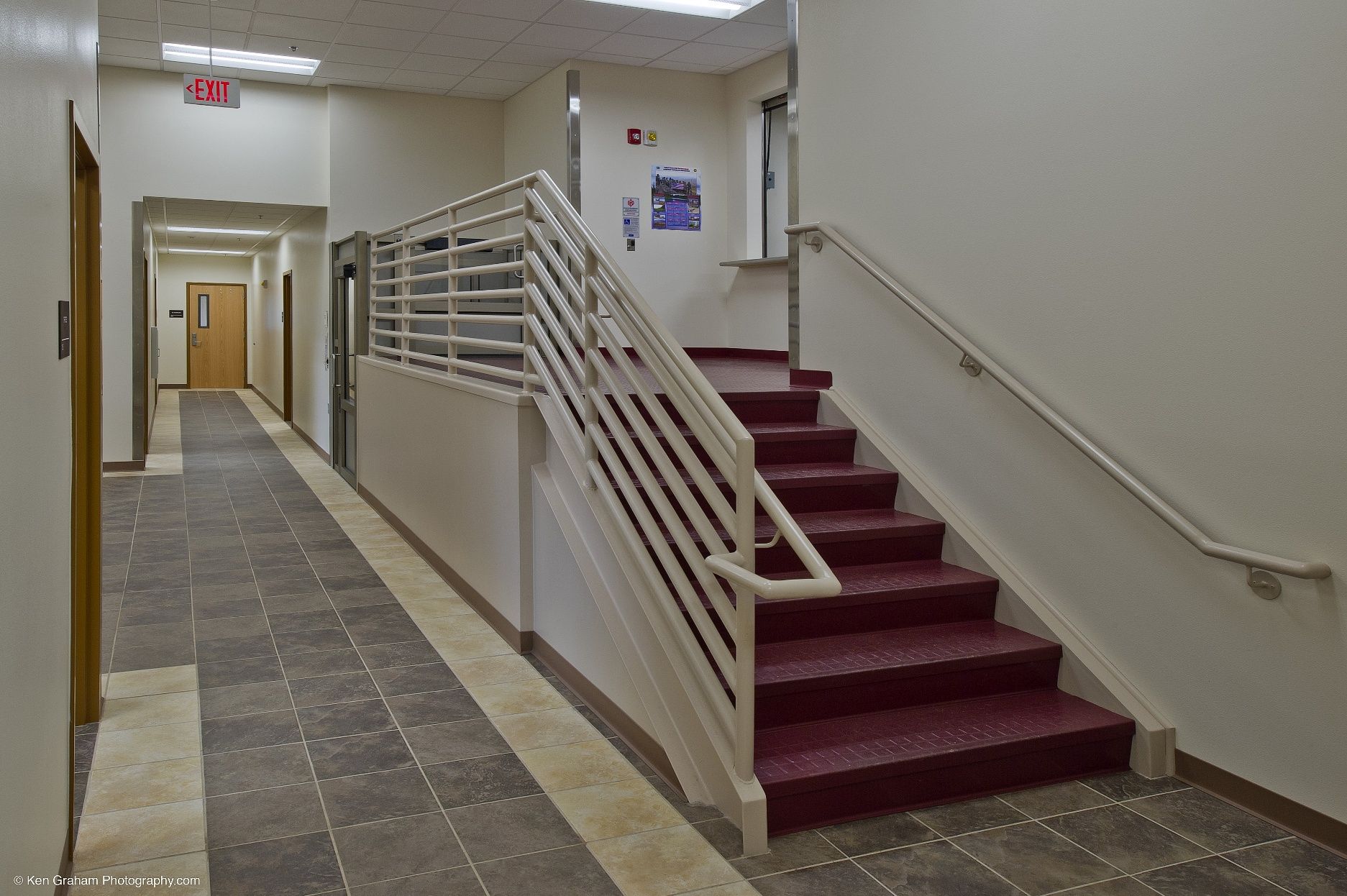 Hallway with burgundy stairs, metal railing, and elevator. Doorway on the left.