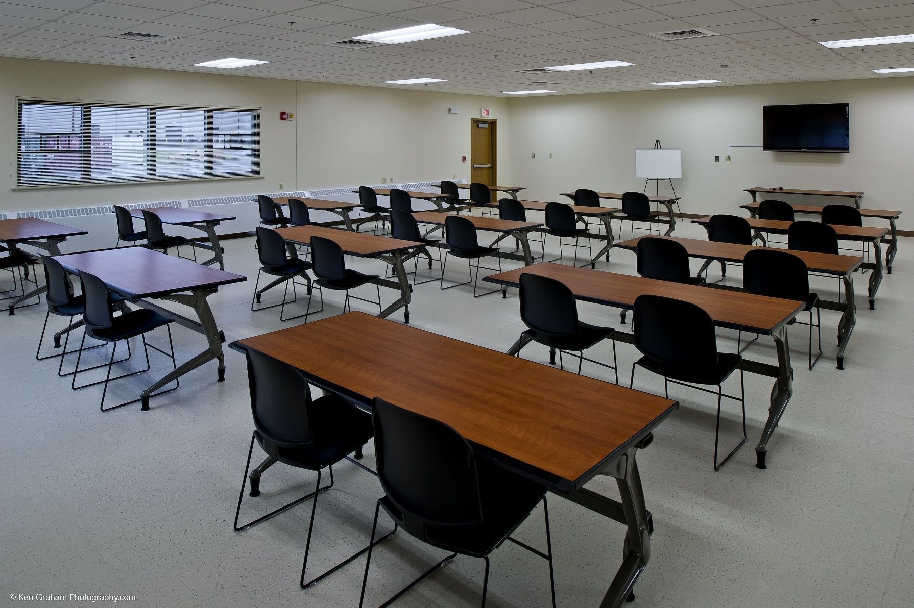 Classroom with rows of tables and black chairs; windows on the left, a TV on the right.