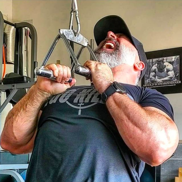 Man with beard gripping pull-up bar, straining to pull, wearing a hat and a black shirt in a gym.