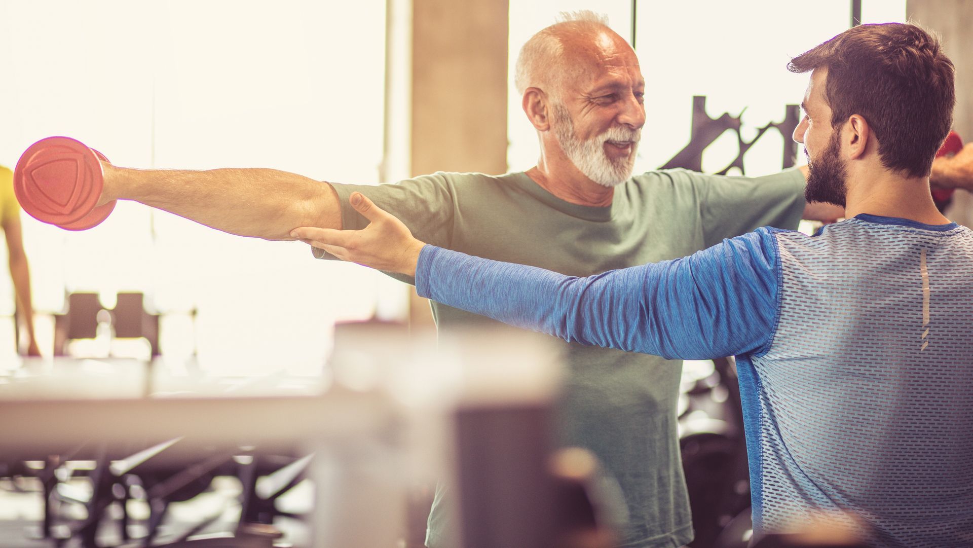 A man is helping another man lift a dumbbell in a gym.