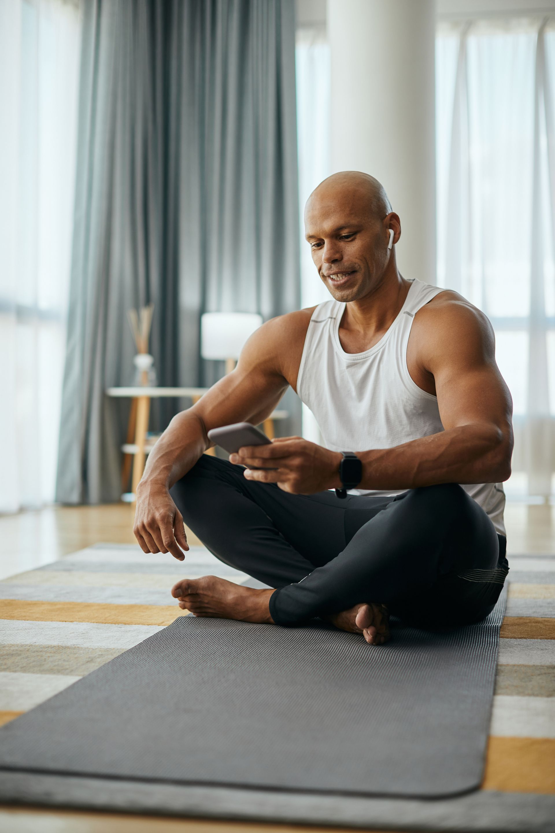 Man sitting cross-legged on yoga mat, looking at phone, wearing earbuds, in a bright room.