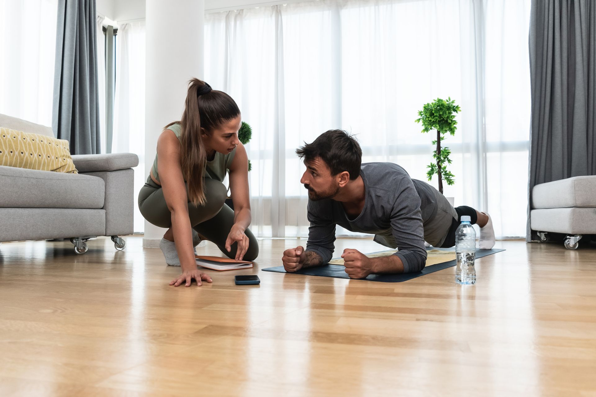 Woman coaching a man doing a plank exercise on a mat indoors.